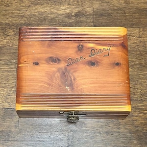 May include: A rectangular wooden box with a natural grain pattern and visible knots. The lid features horizontal grooved lines at the top and bottom edges and the words Dear Diary engraved in script. A small metal latch is centered on the front.
