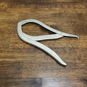 Puede incluir: Un cascanueces plateado con un diseño curvo. El cascanueces tiene un interior dentado y el texto "MIGHTY CRACKER" es visible. La herramienta está abierta y descansa sobre una superficie de madera.