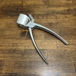 May include: A silver-colored bottle capper tool with a cylindrical head and curved handles. The tool is designed to crimp bottle caps onto glass bottles. The capper is made of metal and sits on a wooden surface.