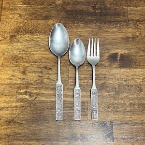 May include: Three silver utensils: a large spoon, a smaller spoon, and a fork. The handles feature a detailed, geometric design. The utensils are arranged on a wooden surface, creating a classic and elegant aesthetic.