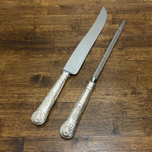 May include: A silver carving knife and sharpening steel set. The knife has a long, curved blade and a decorative handle. The sharpening steel is cylindrical with a matching handle. Both have ornate, shell-like designs. The set is displayed on a wooden surface.