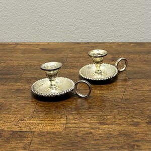 May include: Two silver-coloured candle holders with a small handle on the side. The candle holders have a round base with a beaded edge and a small cup to hold the candle. The candle holders are on a wooden surface.