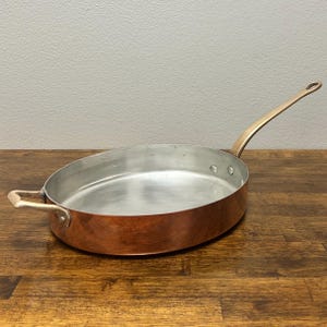 May include: Oval copper-bottomed pan with a long, curved handle and a smaller handle on the opposite side. The interior is silver-colored, and the exterior is a warm copper hue. The pan sits on a wooden surface.