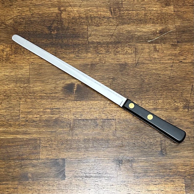 Puede incluir: Un cuchillo de pan largo de acero inoxidable con mango negro. El mango est&aacute; asegurado con dos remaches dorados. El cuchillo est&aacute; colocado en diagonal sobre una superficie de madera.