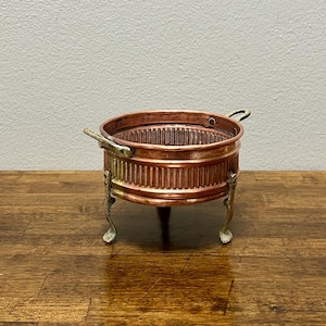 May include: A small, copper-colored metal bowl with a ribbed exterior and two small handles. The bowl sits on three ornate, brass-colored legs. The interior of the bowl is visible and has a textured surface.