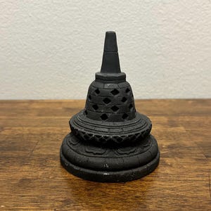 Può includere: Una scultura a forma di stupa color nero opaco, caratterizzata da una base a gradini, una cupola centrale con intagli a forma di diamante e una punta conica. L'oggetto presenta una superficie ruvida simile alla pietra con rilievi.