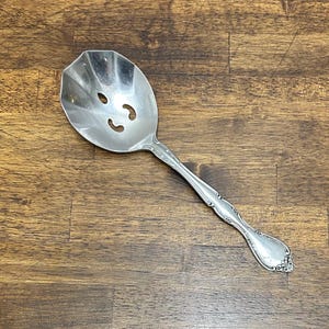 Op de afbeelding: Een zilveren serveerlepel met een uitgesneden kom en een sierlijk handvat. De lepel heeft drie gebogen sleuven in de kom en een decoratief bloemmotief aan het uiteinde van het handvat. De lepel is gemaakt van glanzend metaal.