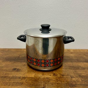 May include: Stainless steel cooking pot with lid. The pot has black handles and a black knob on the lid. The pot is decorated with a band of orange and red geometric shapes around the base. The pot is sitting on a wooden surface.