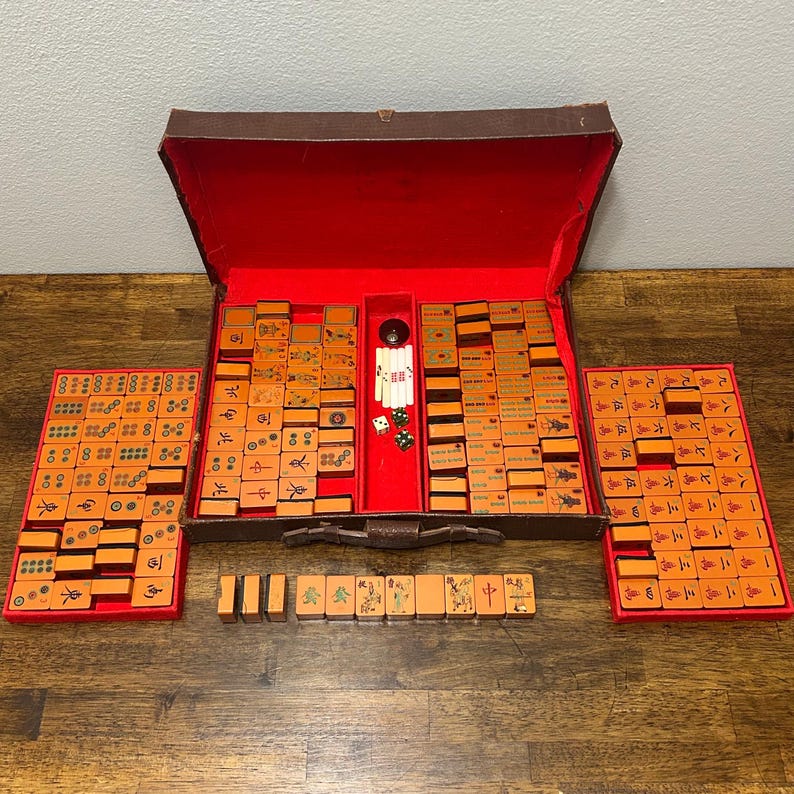 May include: A vintage Mahjong set featuring orange tiles with black and green engraved symbols, housed in a brown leather-like case with a bright red interior lining. The set includes two matching red trays, white betting sticks, and two small dice.