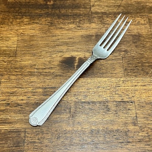 May include: A silver-toned metal dinner fork featuring a four-tine head and a slender handle. The handle is decorated with a beaded border and a shell-like motif at the base, with an embossed scroll pattern near the neck of the fork.