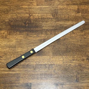 Puede incluir: Un cuchillo de pan largo de acero inoxidable con mango negro. El mango tiene dos remaches dorados. El cuchillo est&aacute; en &aacute;ngulo sobre una superficie de madera.