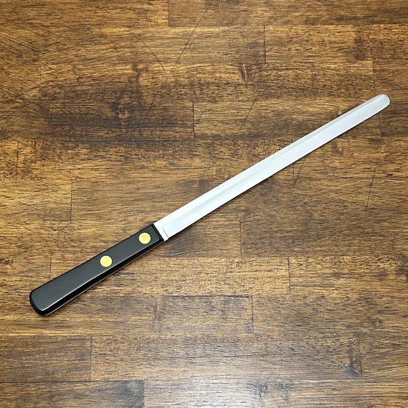 Puede incluir: Un cuchillo de pan largo de acero inoxidable con mango negro. El mango tiene dos remaches dorados. El cuchillo est&aacute; en &aacute;ngulo sobre una superficie de madera.
