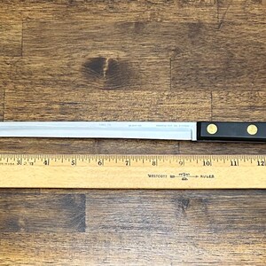 Puede incluir: Un cuchillo para rebanar largo de acero inoxidable con mango negro y dos remaches dorados. El cuchillo mide aproximadamente 35,5 cm de largo, con una hoja recta. Una regla de madera se coloca debajo.