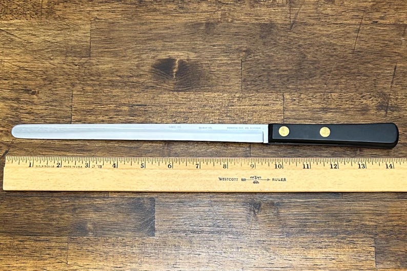 Puede incluir: Un cuchillo para rebanar largo de acero inoxidable con mango negro y dos remaches dorados. El cuchillo mide aproximadamente 35,5 cm de largo, con una hoja recta. Una regla de madera se coloca debajo.