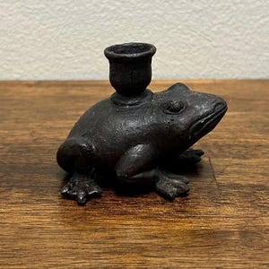 May include: A dark brown, textured cast metal candle holder shaped like a frog. The frog is depicted in a seated position with a small, cylindrical candle cup centered on its back. The surface of the metal has a matte, weathered appearance.