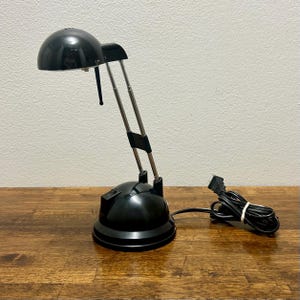 May include: A black desk lamp with a dome-shaped shade and a dual-arm adjustable metal stand. The base is circular and features a black power switch. A black electrical cord with a standard plug is coiled next to the lamp on the surface.