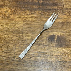 May include: A silver fork with three tines, set against a wooden surface. The handle has a textured, hammered appearance. The fork is designed for serving or eating.