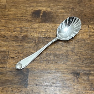 Puede incluir: Una cuchara de servir plateada con un cuenco en forma de concha y un mango largo y delgado. La cuchara está hecha de un metal brillante y reflectante y está diseñada para servir. El mango tiene un diseño decorativo en el extremo.