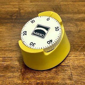 May include: A yellow kitchen timer with a white circular dial featuring black numbers from 0 to 50. The center of the dial displays a black square logo with the white text MIRRO and a curved white line above the brand name.