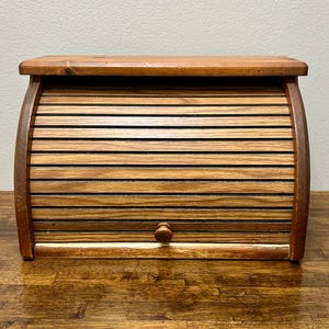 May include: Wooden bread box with a roll-top door. The box is made of light brown wood with a darker brown trim and a small wooden knob. The roll-top door is made of horizontal wooden slats. The bread box is sitting on a wooden surface.