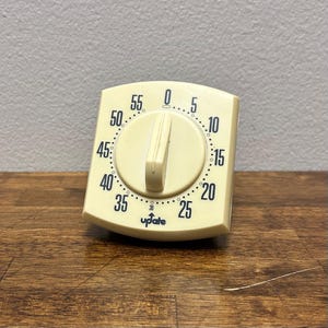 May include: A vintage, off-white kitchen timer with a dial and a large, central knob. The timer has black numerals marking the minutes, from 0 to 55, and the word "update" is printed at the bottom. The timer is set on a wooden surface.