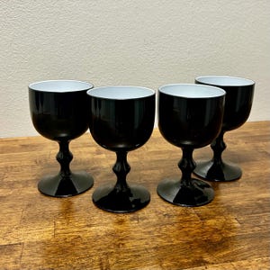 May include: Four black wine glasses with white interiors are arranged on a wooden surface. The glasses have a rounded bowl shape, a slender stem, and a circular base. The glasses are evenly spaced.