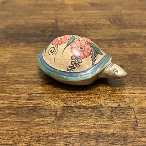 May include: A ceramic turtle figurine. The beige shell is decorated with red flowers and green leaves, and outlined with a blue and green border. The turtle's head is a light beige colour.