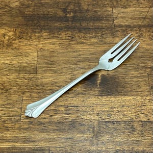 May include: A silver fork with four tines and a decorative handle. The fork is made of a shiny metal and is set against a wooden background. The handle has a detailed, curved design.