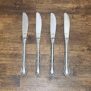 May include: Four silver-colored stainless steel dinner knives arranged vertically. Each knife features a rounded blade and a decorative handle with embossed scrollwork details. The blades are stamped with the text Stainless Steel Japan.