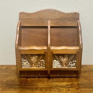 May include: A wooden wall-mounted organizer with two compartments, each featuring a clear window filled with decorative dried plants and seeds. The organizer has a curved top and six small hooks below.