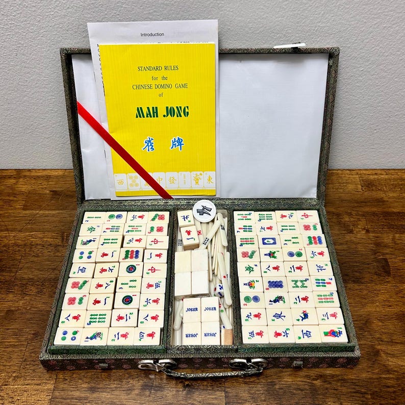 May include: Open mahjong set with ivory tiles, a yellow rule book, and a red ribbon. The set includes tiles with various symbols and characters, and the text "MAH JONG" is visible on the book. The set is in a decorative case.