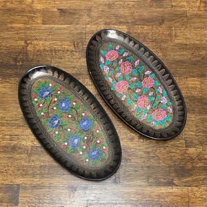 May include: Two oval-shaped black trays featuring intricate floral patterns. One tray displays blue flowers with green leaves, while the other features pink flowers with green leaves. Both trays have a textured gold-toned center and black borders.