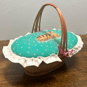 Puede incluir: Una cesta ovalada de mimbre con un asa alta arqueada y una tapa acolchada. La tapa est&aacute; cubierta de tela color verde azulado con un peque&ntilde;o patr&oacute;n floral blanco y un gato atigrado marr&oacute;n bordado en punto de cruz. Un volante blanco bordea la tapa.