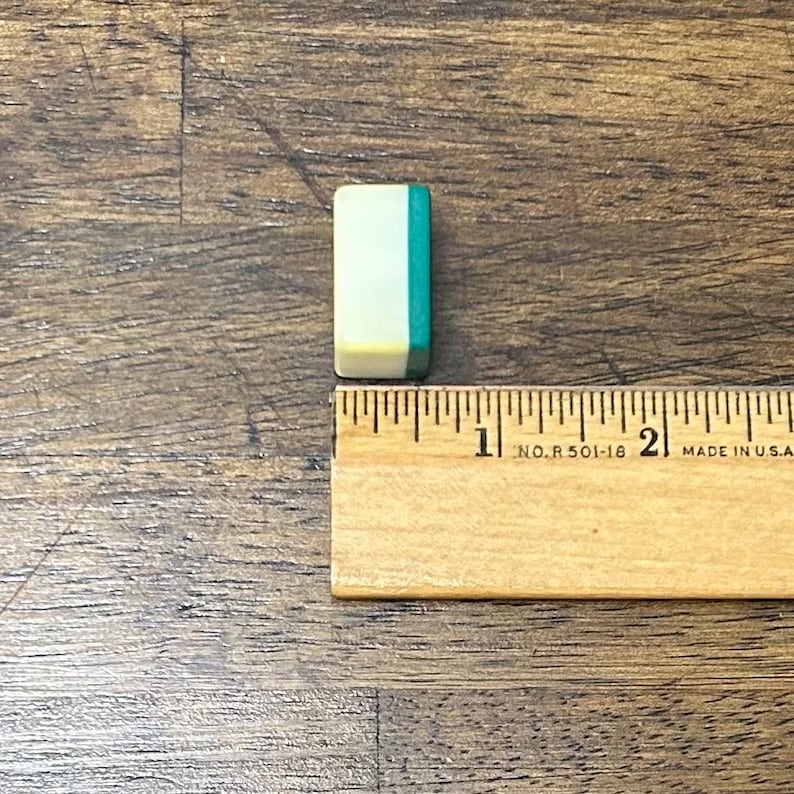 May include: A small, rectangular game tile with a cream-colored center, a teal side, and a yellow edge. The tile is approximately 0.75 inches long. A wooden ruler is next to the tile.