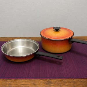 Puede incluir: Una sartén de acero inoxidable y una olla cubierta con asas negras. La batería de cocina tiene un exterior degradado naranja y amarillo. El juego está sobre un mantel individual morado.
