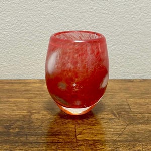 May include: A stemless, round glass with a vibrant red and white marbled design. The glass has a thick base and a smooth, rounded shape. The glass is empty and sits on a wooden surface.