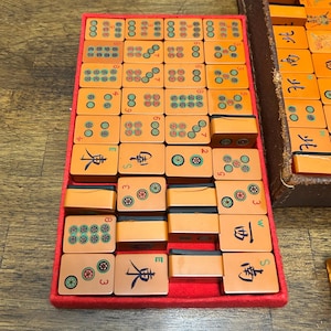 May include: A set of orange Mahjong tiles arranged in a red-lined tray. The tiles feature various green and black circular patterns, Chinese characters, and directional symbols. Some tiles are blank on the face, while others show numbers.