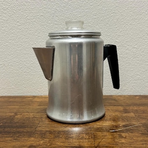 以下が含まれることがあります： 蓋に透明なガラスノブと黒いハンドルが付いた、ヴィンテージのアルミニウム製コーヒーパーコレーター。コーヒーポットには注ぎ口があり、木製の表面に置かれています。パーコレーターは、クラシックなキッチン家電です。