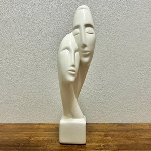 May include: A tall, off-white ceramic sculpture featuring two stylized, elongated faces positioned one slightly behind the other. The smooth, glossy finish highlights the abstract facial features, including closed eyes and defined lips, all resting on a square base.