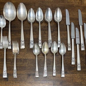 Puede incluir: Una colección de cubiertos antiguos, que incluye cucharas, tenedores y cuchillos, dispuestos sobre una superficie de madera. La cubertería plateada presenta detalles florales decorativos en los mangos. La disposición incluye varios tamaños de cucharas y cuchillos.