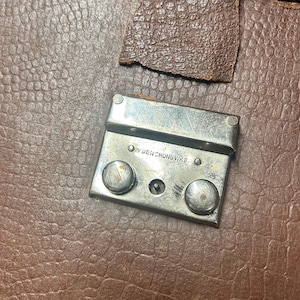 May include: A brown textured leather surface featuring a rectangular silver-toned metal latch mechanism. The metal hardware includes two circular buttons and is engraved with the text YUEN CHUNG WKS. A small piece of brown leather is attached above the latch.