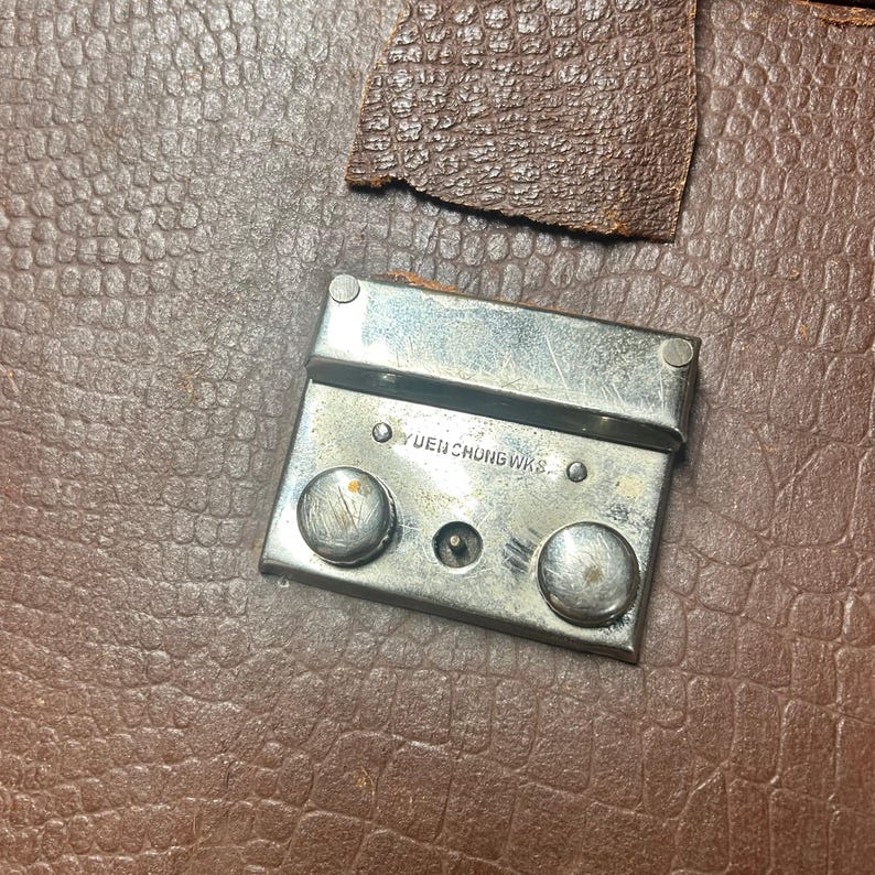 May include: A brown textured leather surface featuring a rectangular silver-toned metal latch mechanism. The metal hardware includes two circular buttons and is engraved with the text YUEN CHUNG WKS. A small piece of brown leather is attached above the latch.
