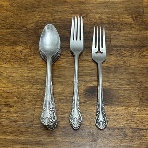 May include: A collection of vintage silverware, including spoons and forks, arranged on a wooden surface. The silverware has ornate, detailed handles and a polished, reflective finish, suggesting a classic or antique style.