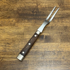 May include: A stainless steel carving fork with a dark brown wooden handle. The handle is secured with silver-colored metal bands and rivets. The fork has two tines and is designed for handling cooked meats.
