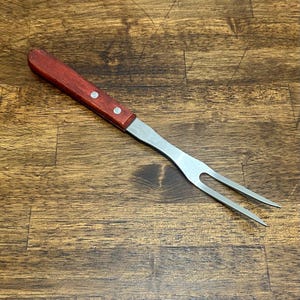 Può includere: Un forchettone da carne a due rebbi con un manico in legno liscio color marrone rossiccio, fissato con due rivetti in metallo color argento. La forchetta presenta una testa in acciaio inossidabile lucido con due rebbi lunghi e affilati.