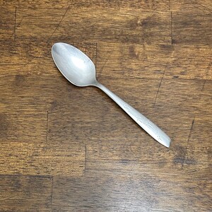 Op de afbeelding: Een zilveren lepel met een afgeronde kom en een lange steel. De steel heeft een decoratief geëtst ontwerp. De lepel ligt op een donkere houtnerf achtergrond.