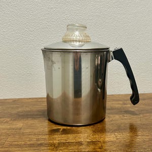 Könnte beinhalten: Eine Perkolator-Kaffeekanne aus Edelstahl mit zylindrischem Körper, schwarzem Kunststoffgriff und einem durchsichtigen Glasknauf auf dem Deckel. Die Metalloberfläche ist glatt und reflektierend, ergänzt durch einen stabilen Seitengriff.