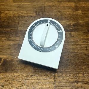 Puede incluir: Un temporizador de cocina blanco con parte superior redondeada y base plana. El centro presenta un dial circular negro con marcas numéricas blancas del 0 al 60, indicando minutos. Una perilla giratoria blanca con un pequeño puntero está en el centro.