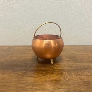 May include: A small, copper-colored cauldron with a brass handle and three brass feet. The round pot has a polished finish and is likely used for decorative purposes. The cauldron sits on a wooden surface.