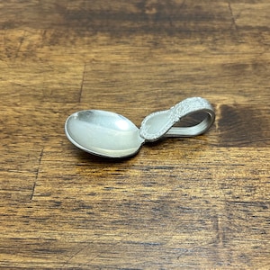 May include: A small, silver spoon with a decorative handle. The spoon's bowl is oval-shaped, and the handle curves into a loop. The handle features an ornate, embossed design. The spoon is likely for serving or decorative purposes.